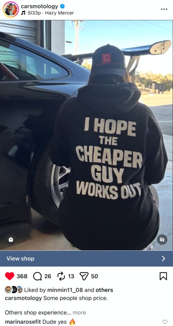 Auto detailer wearing a Carsmotology hoodie working next to a performance car inside a detailing shop.