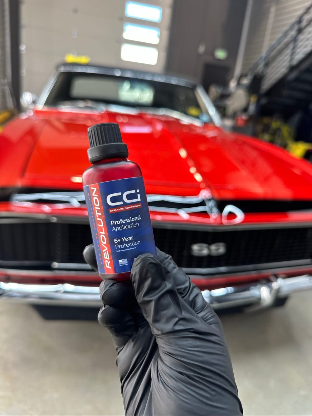 Bottle of CCi Revolution ceramic coating held in front of a red 1967 Camaro SS during a professional ceramic coating application