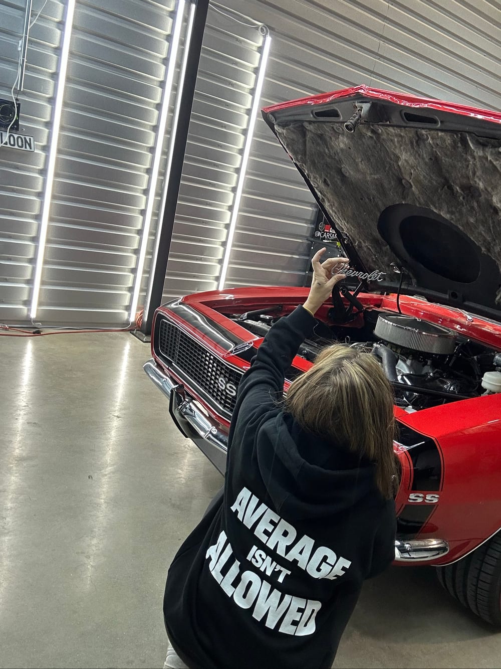Carsmotology technician opening the hood of a red 1967 Camaro SS to detail the engine bay built by Cruzers Customs