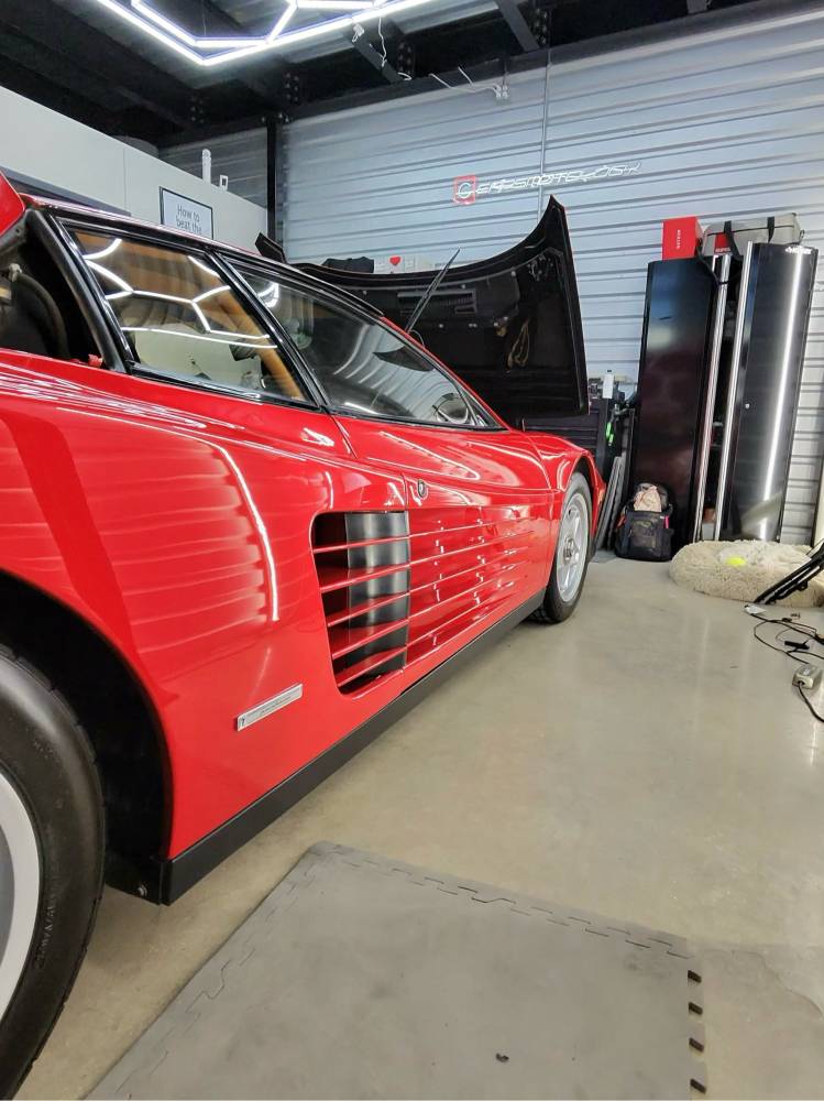 Red Ferrari Testarossa inside the Carsmotology detailing studio with the engine lid open and shop lights reflecting in the side strakes